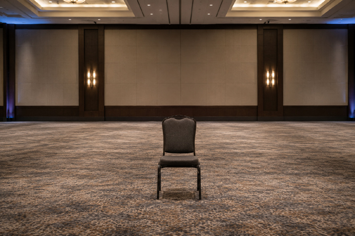 Single empty chair centered in a bare room representing the difference between passive observation and genuine audience engagement at events